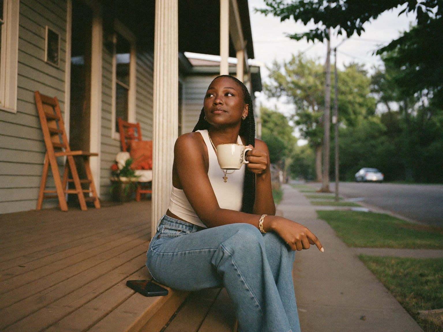 A woman sits on the front porch steps of a home at golden hour, coffee mug in hand, phone on the step beside her